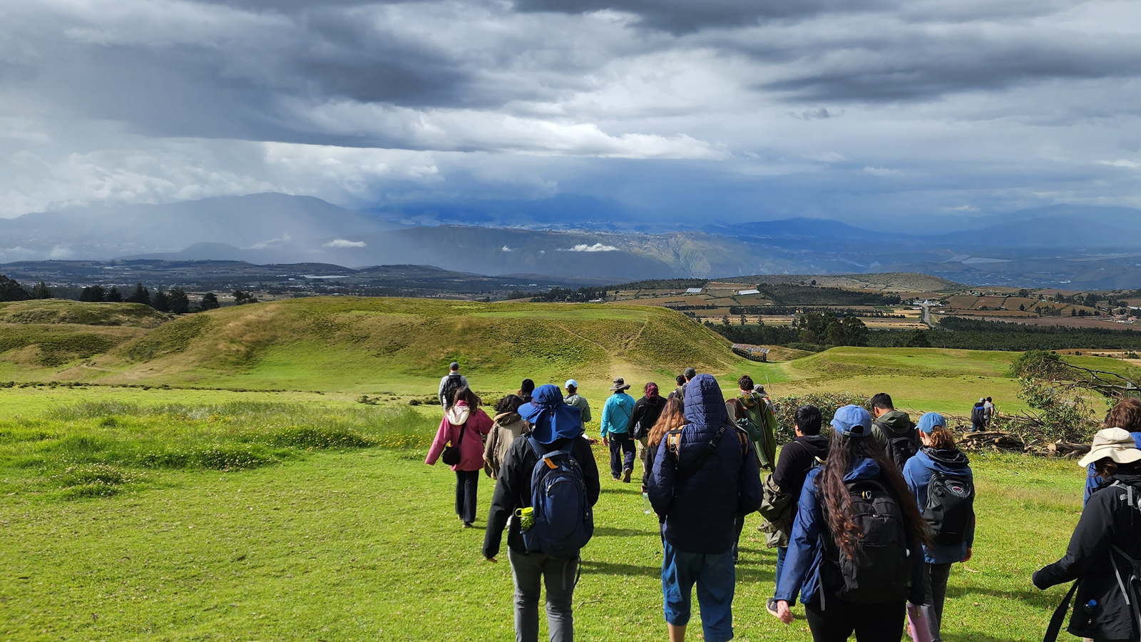 A group of students hikes across rolling green hills under dramatic storm clouds, overlooking a broad valley and distant mountains.