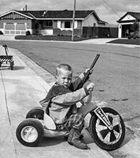 Black and white photo young boy holding a toy gun on a tricyle in front of a suburban house