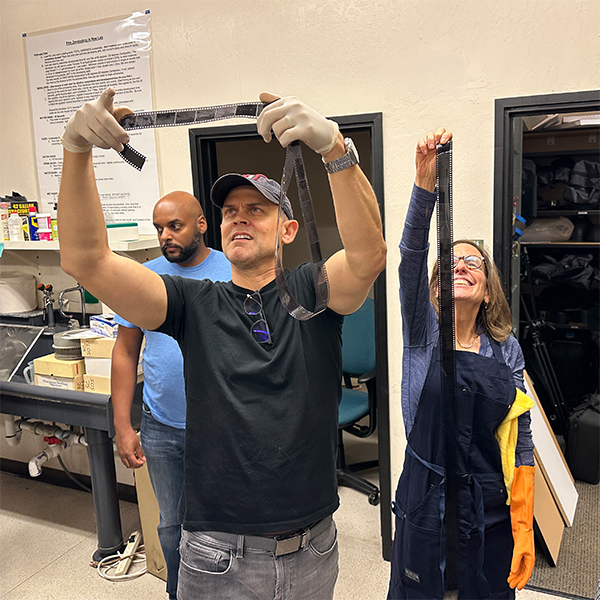 Three people stand in a photography lab examining strips of film. Two individuals wearing gloves carefully inspect negatives while another person observes nearby