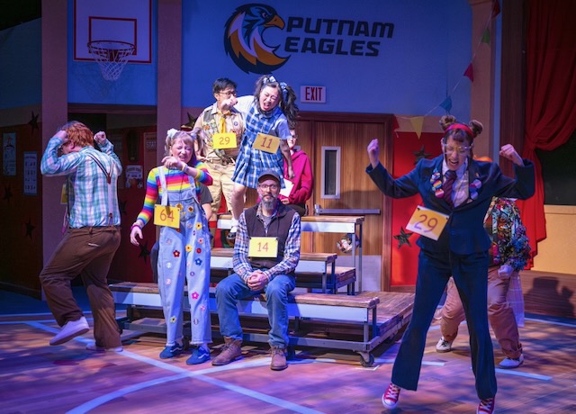 Cast members portraying spelling bee contestants celebrate excitedly on a school gym stage set, wearing numbered badges while one contestant sits calmly on the bleachers.